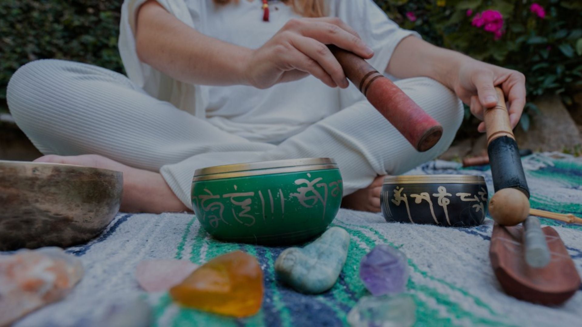 Person sitting outdoors with singing bowls and tools on a colorful mat.